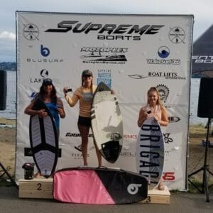Three women standing on a podium with surfboards in front of them, sponsored by Supreme Boats.