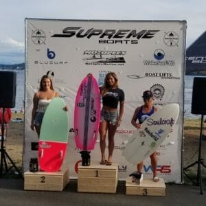 Three women standing on podiums with surfboards in front of them at a Supreme Boats event.
