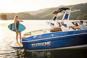 A woman is standing on a Supreme boat holding a surfboard smiling toward her family.