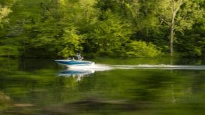 A person is driving a Supreme boat on a lake with trees in the background.