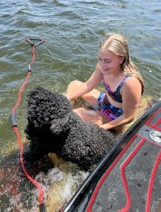 A girl sits on the back of a Supreme boat with a black poodle.