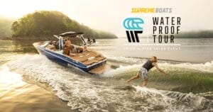 A person wakesurfing behind a speeding boat on a lake with forested hills in the background, under a glowing morning sun. Ad for "The 2024 Supreme Waterproof Tour" on upper right.