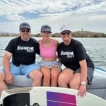 Three people, likely from the Gilmore family, wearing Supreme Boats hats sit together on a boat in open water. Two women in pink and black tops are flanked by a man in a black shirt and blue shorts. A wakeboard rests in front of them, hinting at their Lake Havasu adventure.