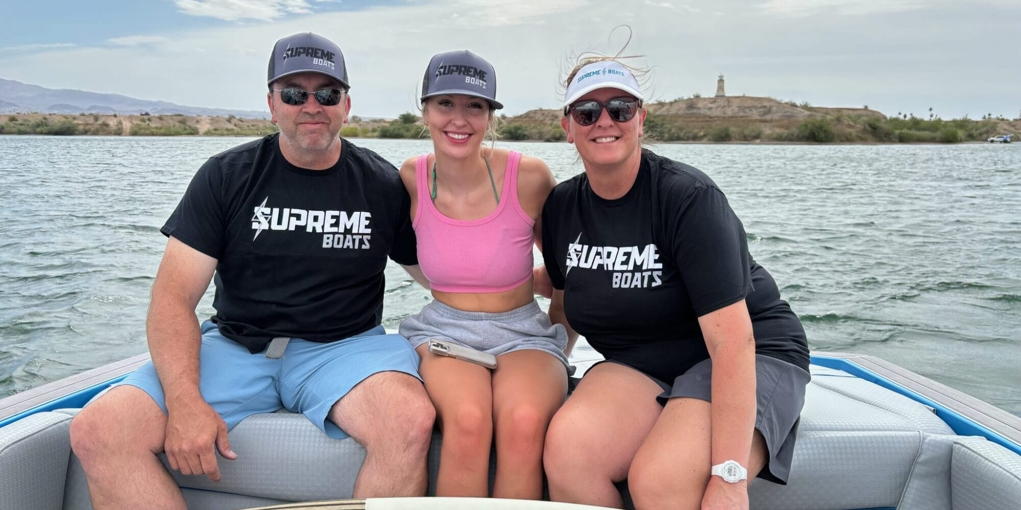Three people, likely from the Gilmore family, wearing Supreme Boats hats sit together on a boat in open water. Two women in pink and black tops are flanked by a man in a black shirt and blue shorts. A wakeboard rests in front of them, hinting at their Lake Havasu adventure.