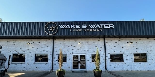 Front exterior of Wake & Water Lake Norman dealership featuring a large sign, double glass doors, brick walls, and planters flanking the entrance. Proudly showcasing Supreme Boats and Centurion models.