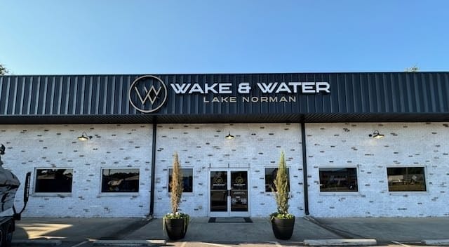 Front exterior of Wake & Water Lake Norman dealership featuring a large sign, double glass doors, brick walls, and planters flanking the entrance. Proudly showcasing Supreme Boats and Centurion models.